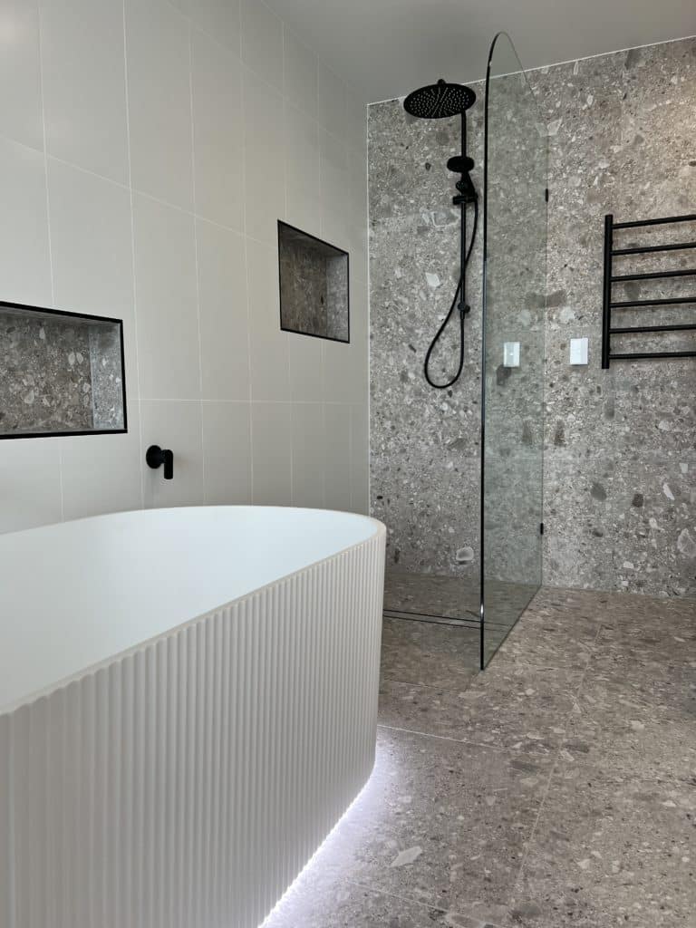 white bathtub in front of shower area that has been tiled with grey terrazzo tiles from floor up to the wall. Terrazzo tiles are contrasted against white 600x600 tiles stacked. Matte black shower rail and features.