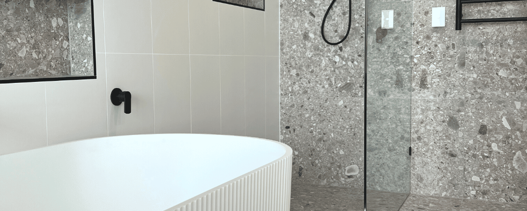 white bathtub in front of shower area that has been tiled with grey terrazzo tiles from floor up to the wall. Terrazzo tiles are contrasted against white 600x600 tiles stacked. Matte black shower rail and features.