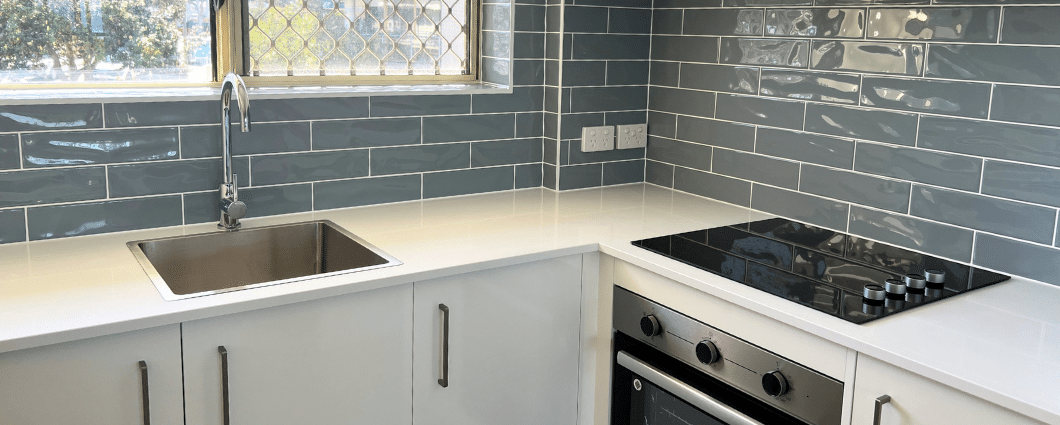 corner view of white kitchen with white stone benchtop and misty grey splashback tiles in a subway pattern.